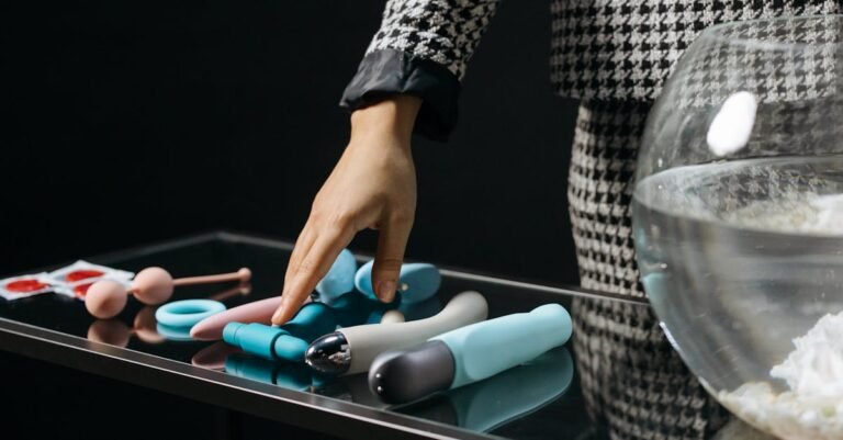 Hand reaching for various colorful silicone adult toys on a reflective glass table. Sensual and vibrant.