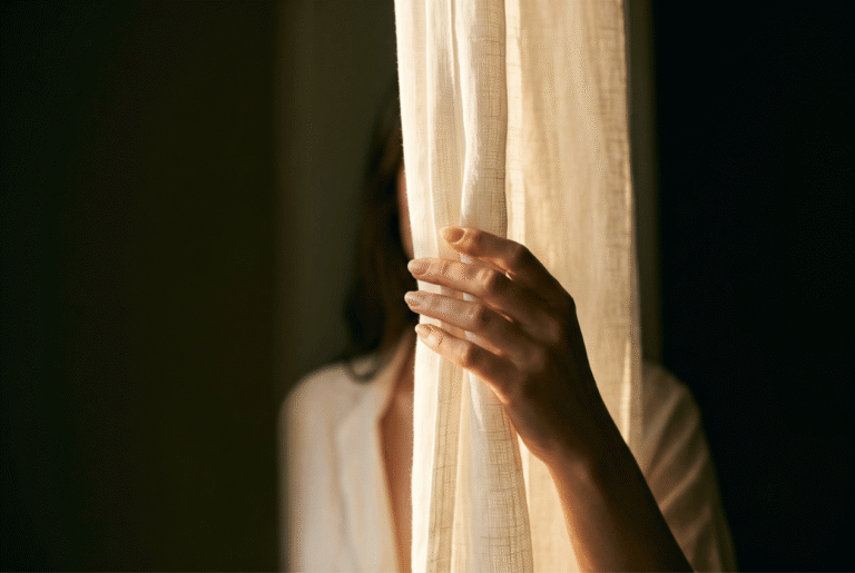 Woman's hand drawing back a sheer curtain toward warm light